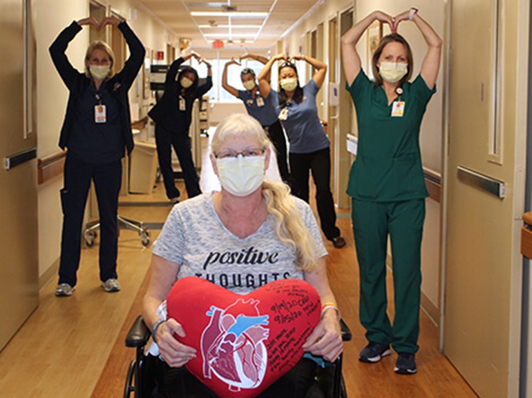 Debra wearing mask and sitting in wheelchair holding red heart-shaped pillow signed by her nurses