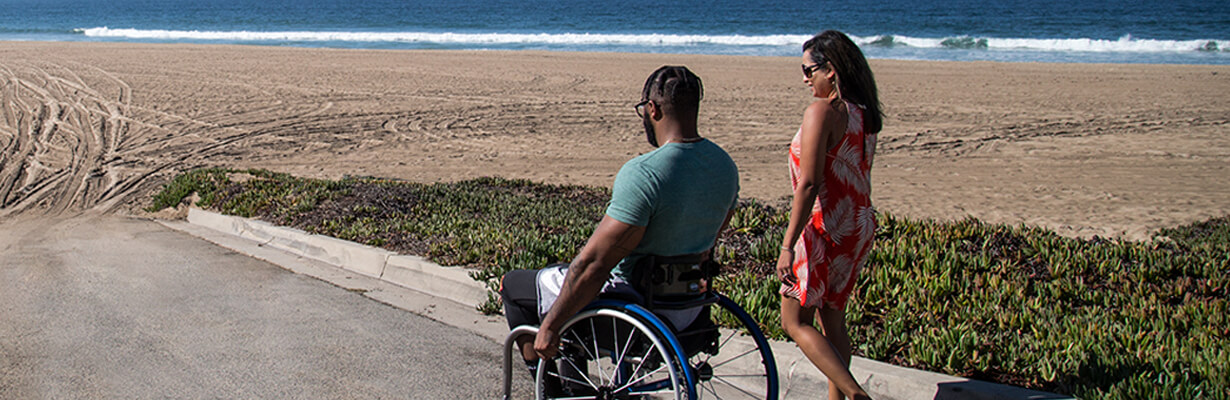 Man in wheelchair and his wife approaching beach and ocean.