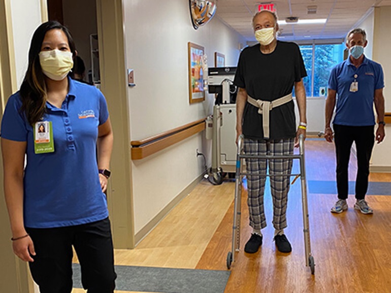 Edward standing with his walker in hospital hallway with two physical therapists